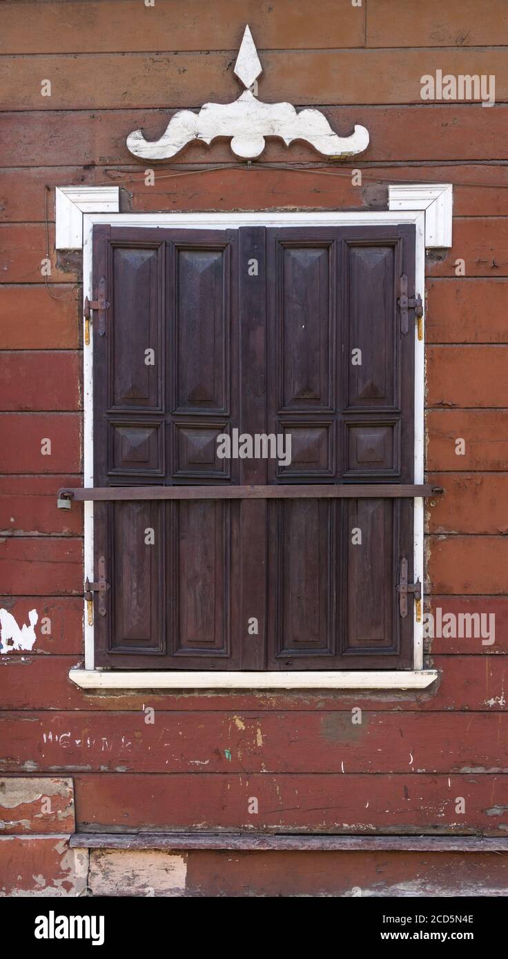 Details der traditionellen Architektur. Braune Holzwand mit Fenster geschlossen mit hölzernen Fensterläden mit Metallbeschlägen und weißen dekorativen Details. Stockfoto