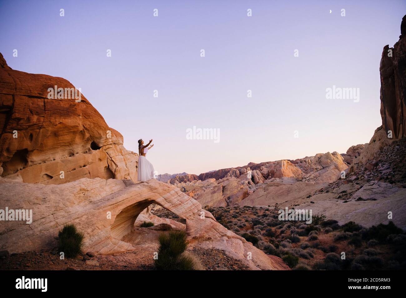 Nymphe-ähnliche Frau in Weiß, die auf einem natürlichen Bogen aus Aztekenstein steht, State Park, Mohave Desert, Overton, Nevada, USA Stockfoto