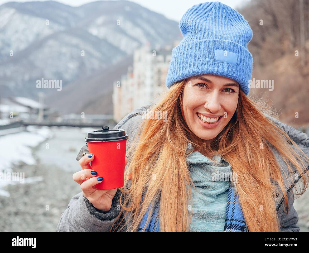 Lächelnd rot Kopf Mädchen hält roten Kaffeeglas mit schwarz Deckel auf verschwommener Berglandschaft Hintergrund Stockfoto
