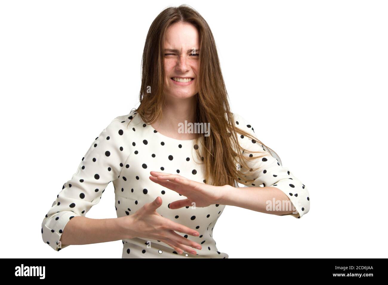 Eine junge Frau mit langen braunen Haaren und einem weißen Polka-Dot-Kleid. Mit einem unzufriedenen, angespannten emotionalen Gesicht. Hält seine Hände horizontal parallel zur Handfläche vor ihm. Isoliert auf Weiß Stockfoto