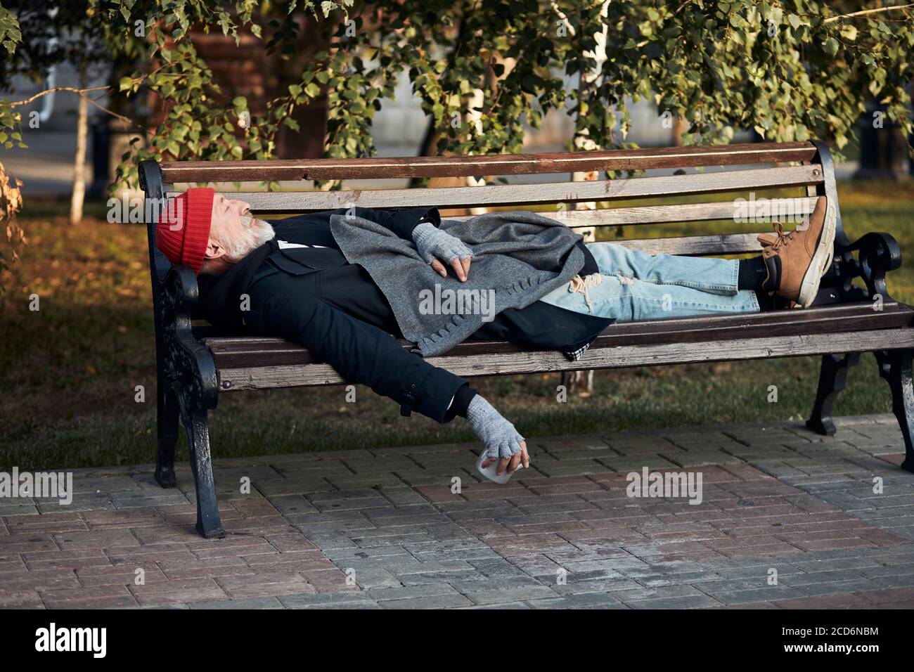 Obdachloser, der tagsüber auf der Bank im Park schläft. Reifer graubärtiger Mann in Straßenkleidung. Seitenansicht Stockfoto