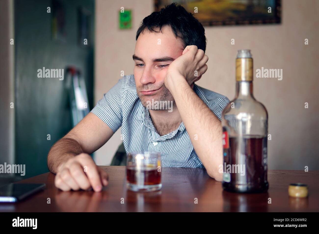 Trauriger, trauriger junger Alkoholiker sitzt mit einer Flasche Rum am Tisch. Das Gesicht eines betrunkenen Mannes. Dumme Emotionen. Depression und Alkoholismus. Stockfoto