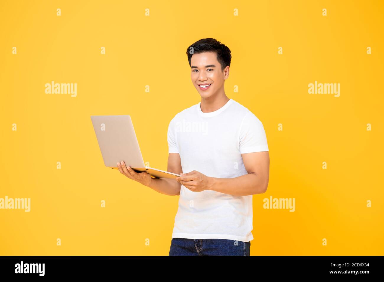 Porträt eines lächelnden jungen schönen asiatischen Mann mit Laptop Computer, während Sie die Kamera in einem isolierten Studio gelben Hintergrund Stockfoto