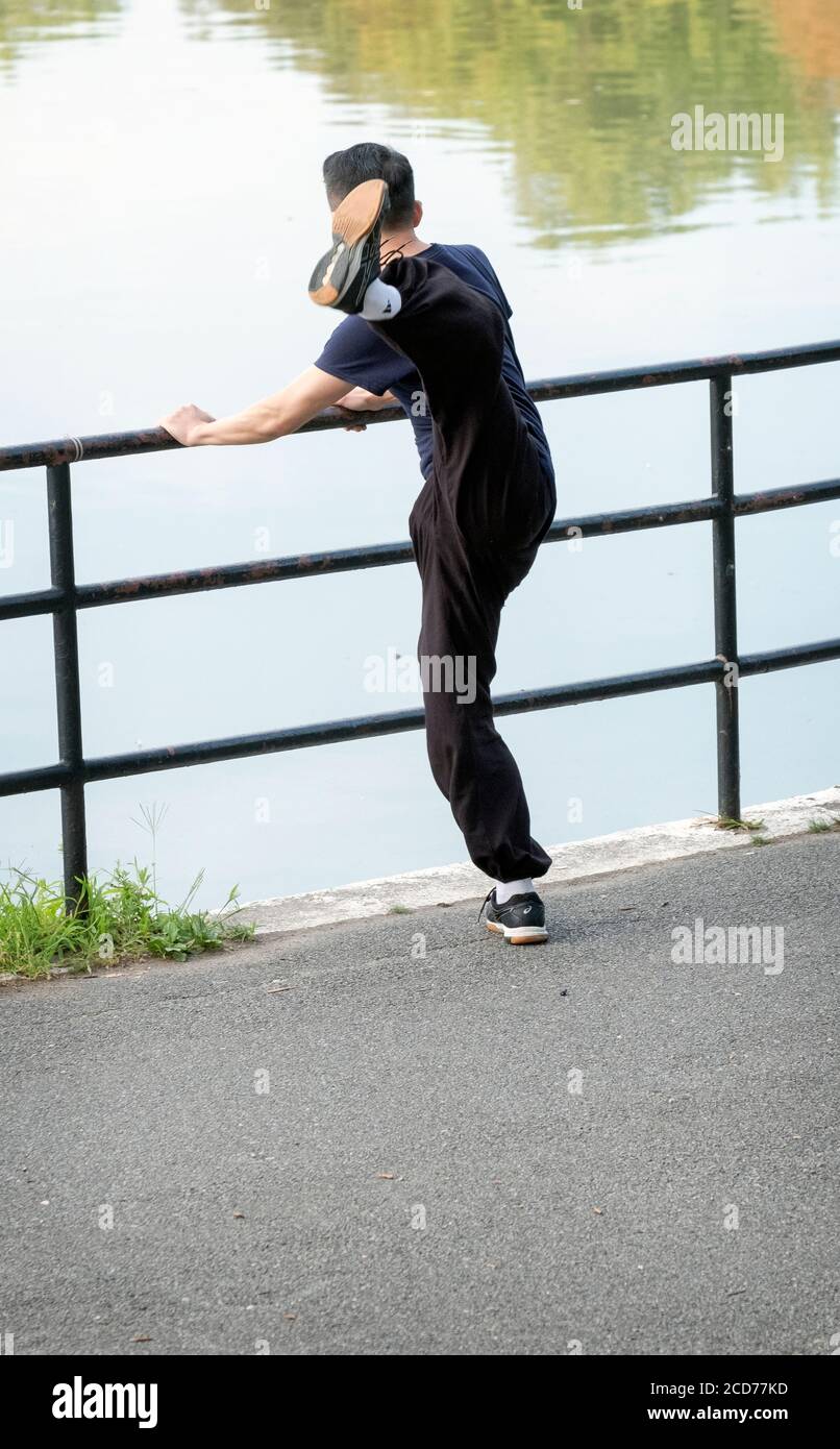 Ein bemerkenswert flexibler asiatischer amerikanischer Mann limbiert sich für Morgenübungen, indem er sein Bein sehr hoch tritt. In Kissena Park, Flushing, Queens, New York. Stockfoto