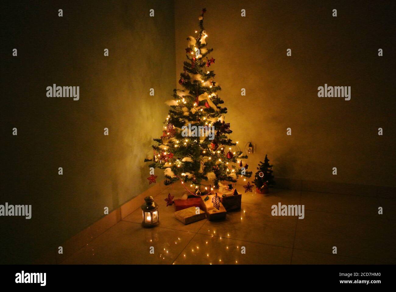 Beleuchteter und dekorierter Weihnachtsbaum auf dunklem Hintergrund mit Geschenken und Laterne darunter. Warme Fadenleuchten erleuchten den Baum. Stockfoto