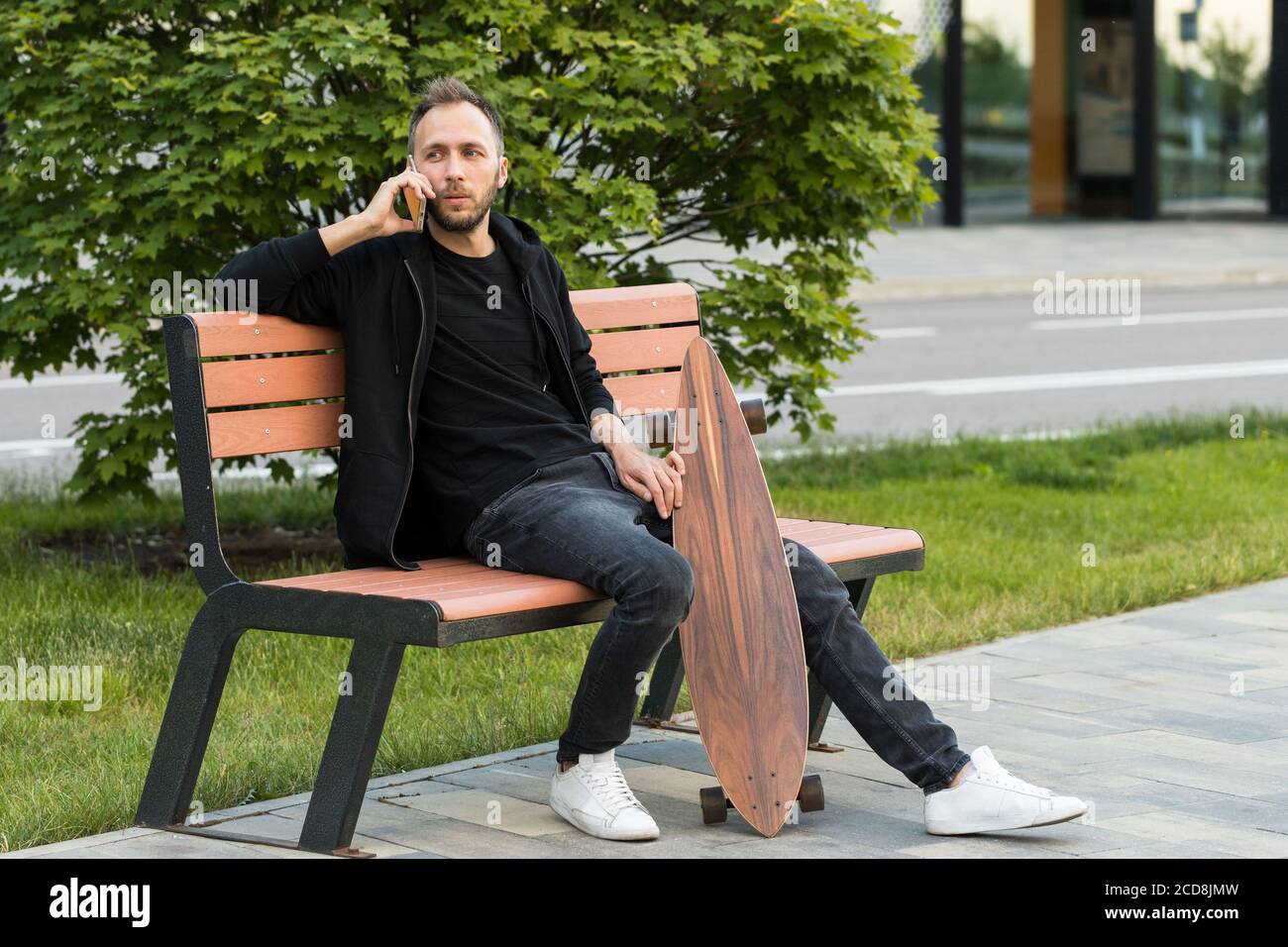 Junger Mann ruht sich aus, sitzt mit Skateboard oder Longboard auf Holzbank, spricht auf Smartphone. Stockfoto