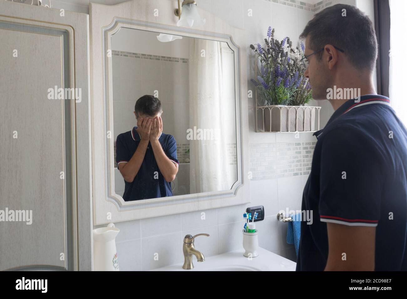 Der junge Mann, der vor dem Spiegel steht, sieht sich in verzweifelter Reflexion mit den Händen im Gesicht. Konzept der Depression oder psychischen Erkrankungen. Stockfoto