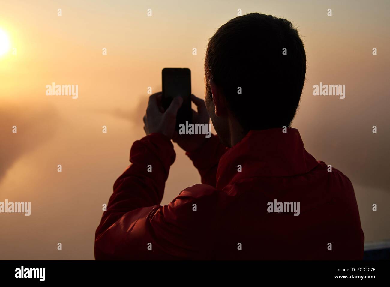Ein Mann auf einer Brücke über den Fluss, der auf dem Handy einen Sonnenaufgang an einem nebligen frühen Morgen fotografiert. Stockfoto
