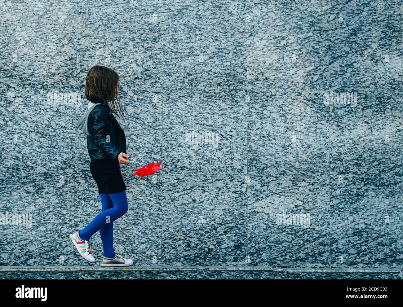 Kleine Stadt stilvolle Mädchen in lässigen Herbst Kleidung zu Fuß entlang graue Granitwand mit rotem Wind Spinner. Leerzeichen für Text. Stockfoto