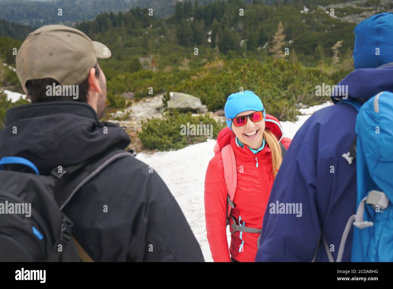 Drei Wanderer tragen Jacken, Berge und Wald teilweise mit Schnee bedeckt, junge Frau in roter Jacke, blaue Kappe und Sonnenbrille Blick auf andere tw Stockfoto