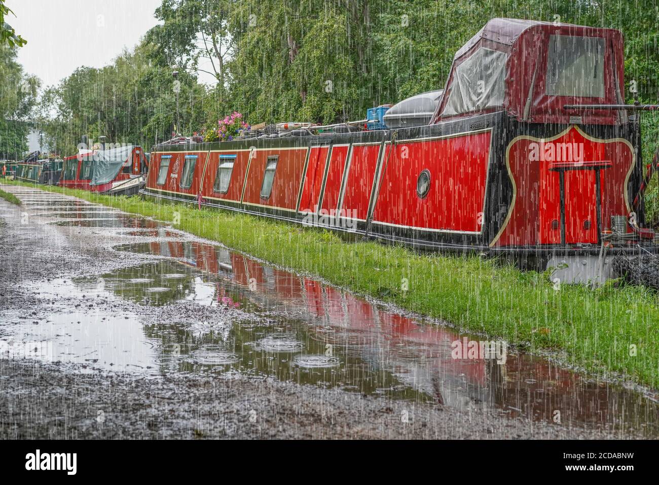 Britische Schmalboote vertäuten entlang eines Kanals Treidelpfad mit Pfützen während der schweren Sommer regen Dusche. Britischer Bootsurlaub im Regen. Stockfoto