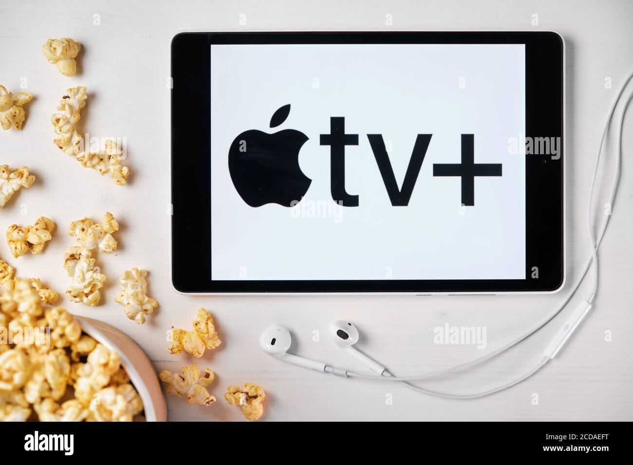 Apple tv plus Logo auf dem Bildschirm des Tablets, das auf dem weißen Tisch lag und darauf Popcorn streute. Apple Ohrhörer in der Nähe des Tablets mit einem Apple Stockfoto