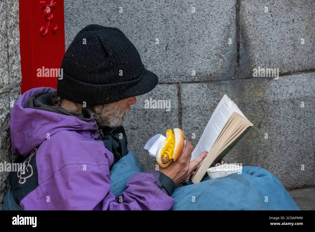 Ein obdachloser Mann wickelte sich in einen alten Schlafsack und aß einen Hot Dog, der ein Buch las, das einen Hut trug, der warm blieb. Stockfoto