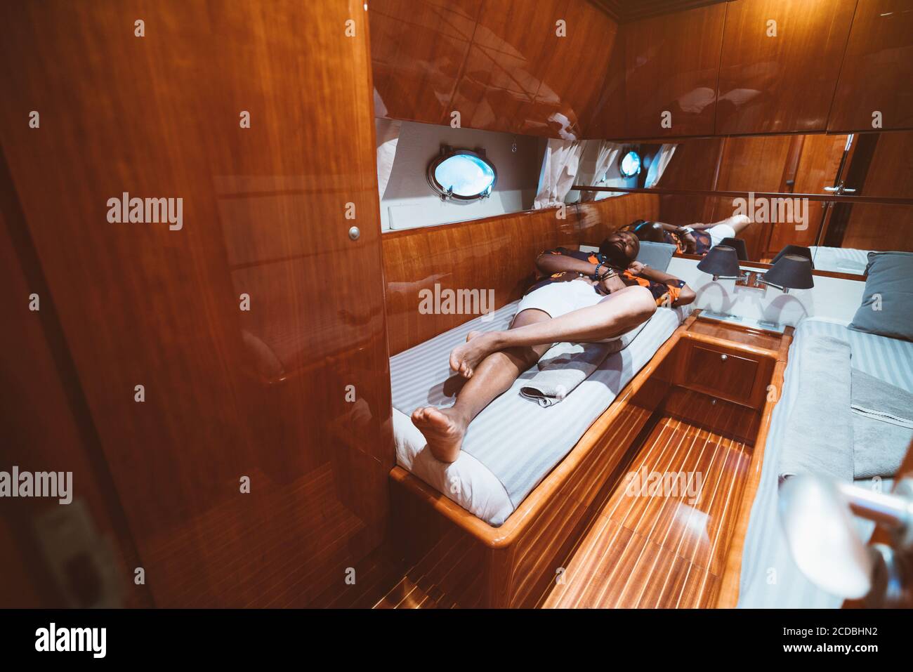 Ein reicher unbeschwerter bärtiger schwarzer Kerl in Shorts liegt auf einem Bett in einer Kabine einer Luxusyacht mit Bullauge, Betten, reflektierenden Holzwänden Stockfoto