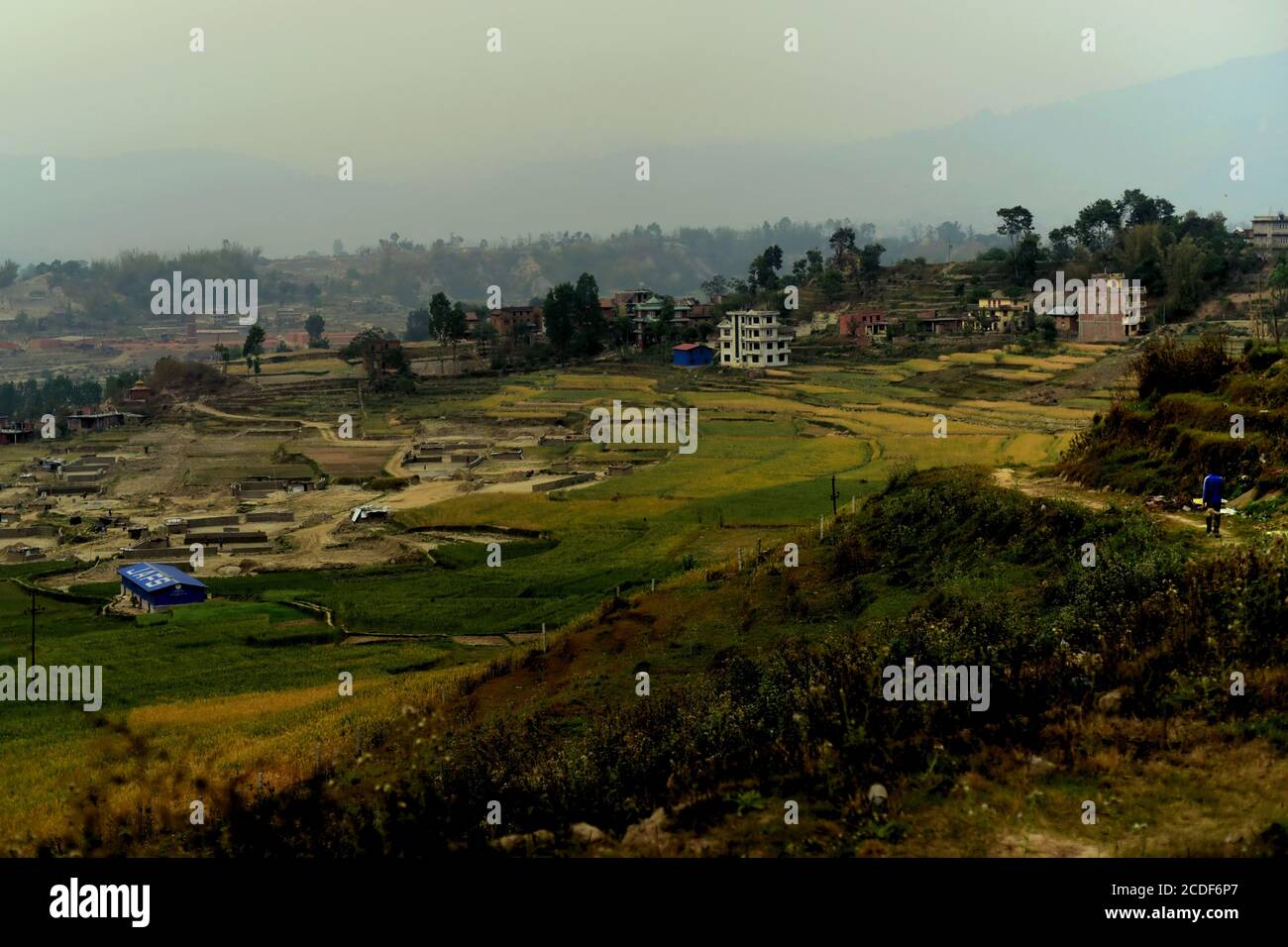 Ein Mann, der auf einem ländlichen Weg geht, mit Blick auf landwirtschaftliche Felder, die teilweise in eine Baustofffabrik umgewandelt wurden, am Stadtrand von Kathmandu, Nepal. Stockfoto