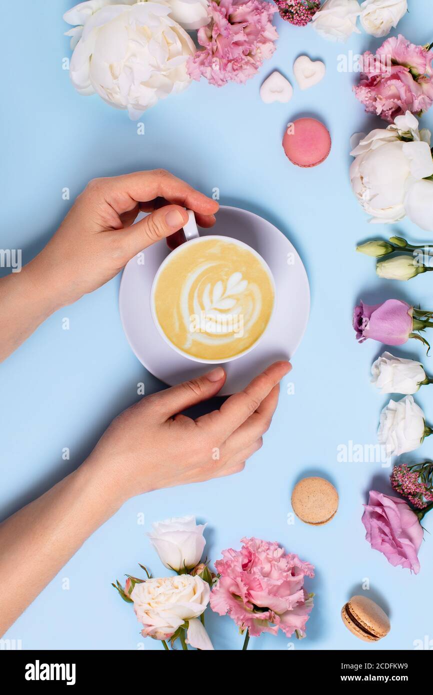 Festliches vertikales Foto mit blauem Hintergrund. Eine Tasse Latte Macchiato in Frauenhänden, umgeben von Rosen, Pfingstrosen, Makronen und Zuckerstücken i Stockfoto