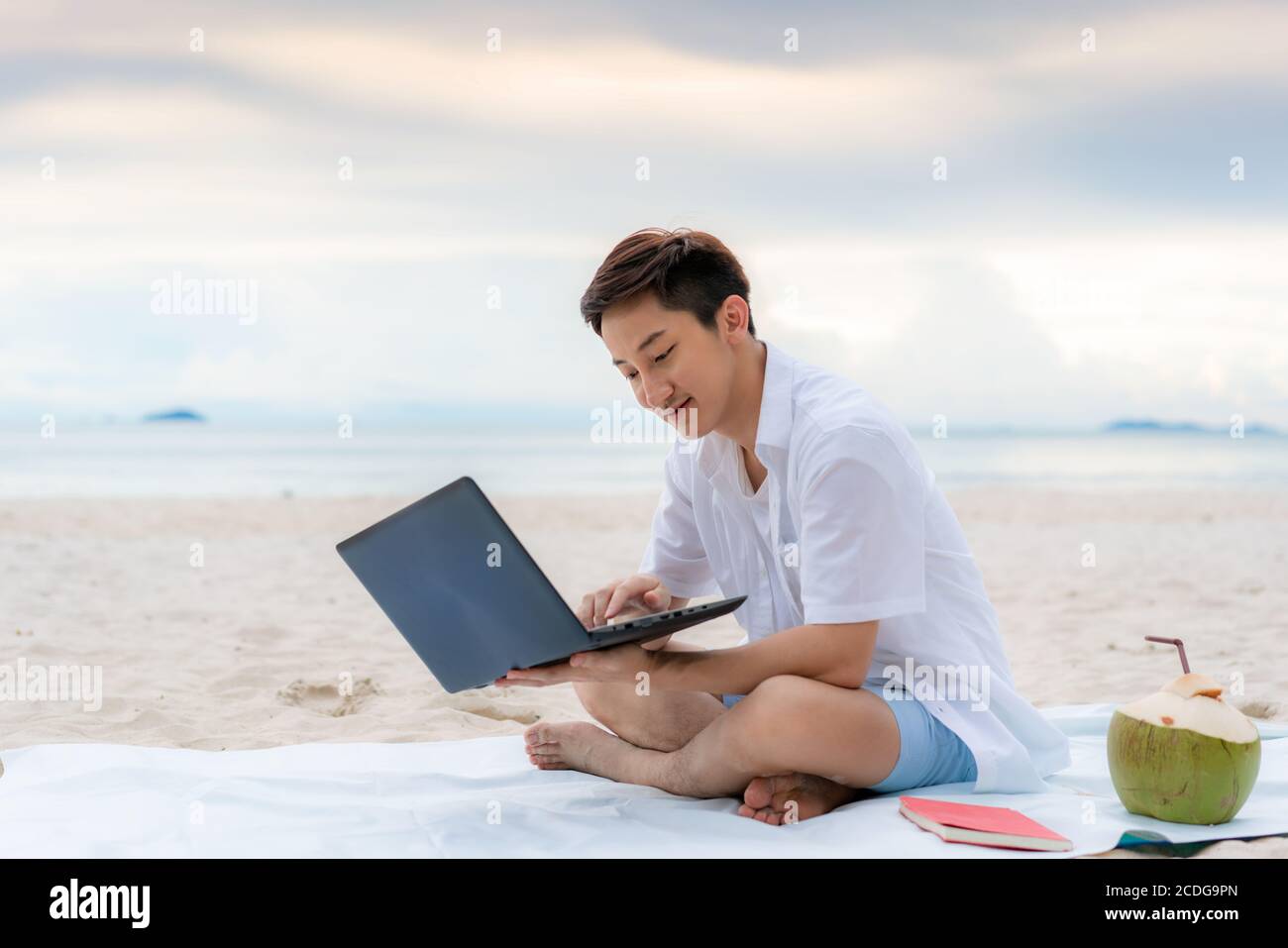 Junger asiatischer Mann, der während seiner Urlaubszeit mit einem Laptop draußen arbeitet, während er am schönen Strand sitzt. Sommer, Urlaub, Urlaub und glückliche Menschen ich Stockfoto Junger asiatischer Mann, der während seiner Urlaubszeit mit einem Laptop draußen arbeitet, während er am schönen Strand sitzt. Sommer, Urlaub, Urlaub und glückliche Menschen ich Stockfoto