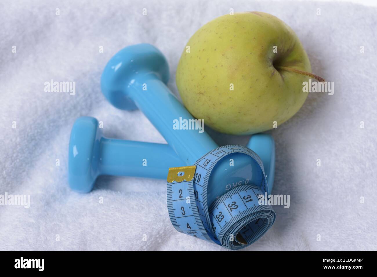 Gesundheits-Regime und Fitness-Symbole. Kurzhanteln nahe grünem Apfel auf weichem hellem Stoffhintergrund. Apfelfrucht und Langhanteln mit blauem Maßband gebunden. Gesundes Lifestyle- und Sportkonzept. Stockfoto