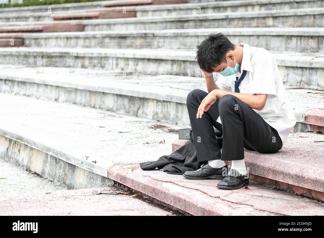 Ausfall arbeitslos gestresst jungen asiatischen Geschäftsmann trägt eine Gesichtsmaske, die Kopf mit Händen. Ein junger Geschäftsmann sitzt verzweifelt auf der Treppe Stockfoto