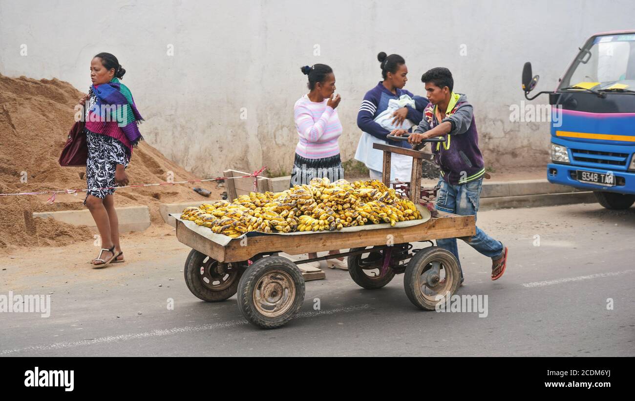 Antananarivo, Madagaskar - 24. April 2019: Unbekannter madagassischer Mann, der Karren mit reifen Bananen auf der Asphaltstraße zieht, andere Frauen und Auto im Backgrou Stockfoto