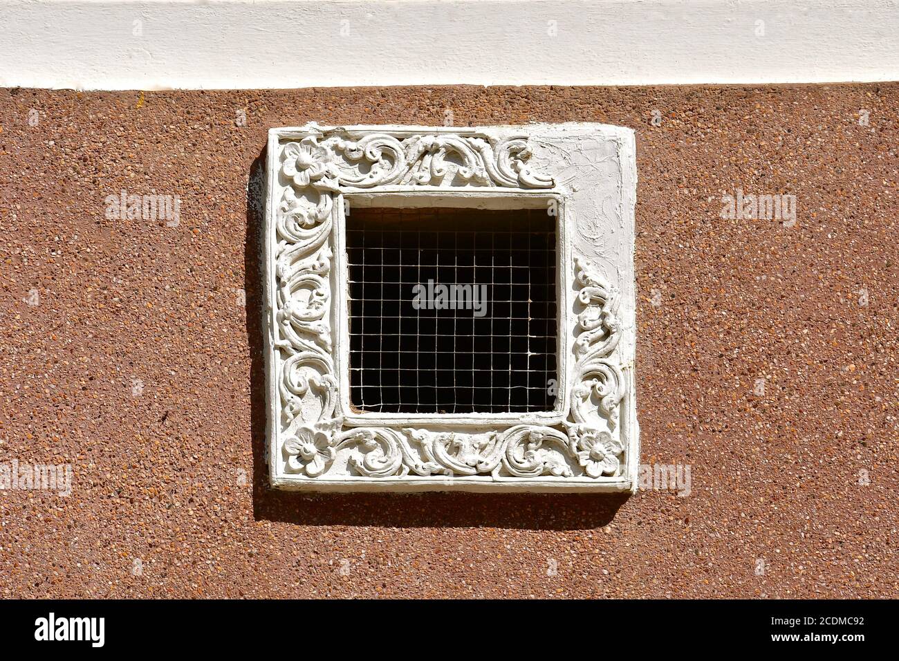 Kleiner quadratischer Lüftungsschlitz mit Hühnerdrahtgitter und einem verzierten weißen Gipsfensterrahmen. Isoliert auf einer sandigen farbigen Kieswand gestrichelt. Stockfoto