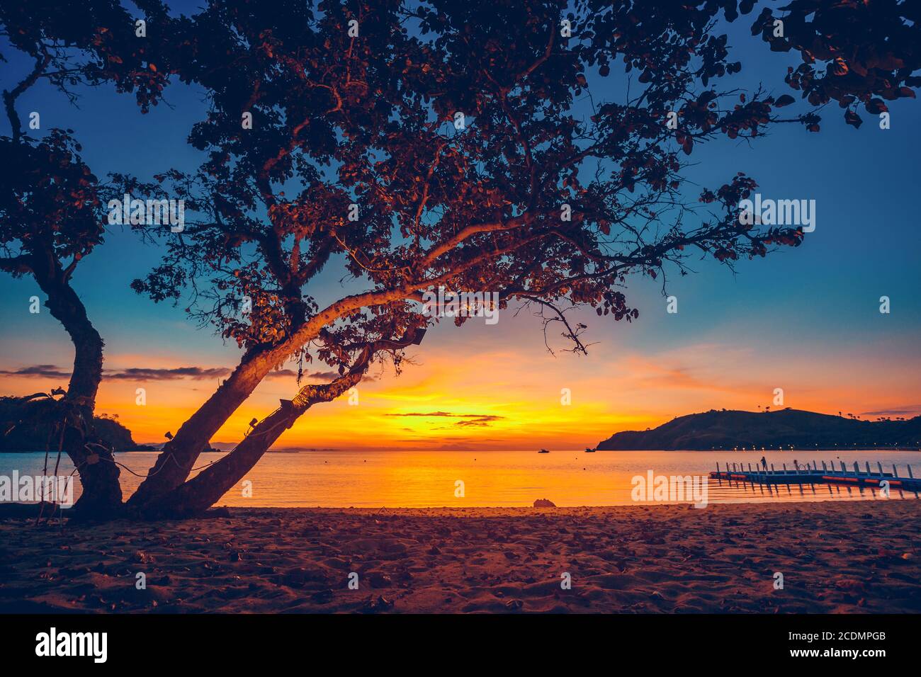 Indonesien Ozean Sonnenuntergang, Baum Silhouette am Sandstrand in der Nähe Hafen. Klippeninsel mit Lichtern Ufer. Schiffe am Meer Bucht Wasser. Klarer Himmel im tropischen Resort. Erstaunliche bunte Naturlandschaft von Asien Stockfoto