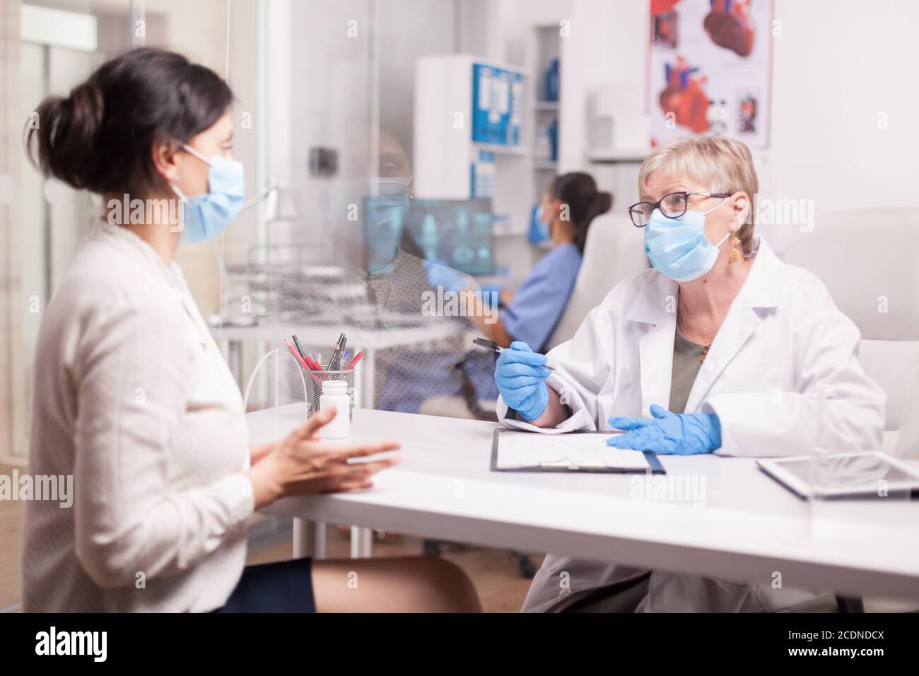 Reifer Arzt mit Gesichtsmaske gegen Coronavirus während der Konsultation mit kranken jungen Frau im Krankenhaus-Büro. Stockfoto