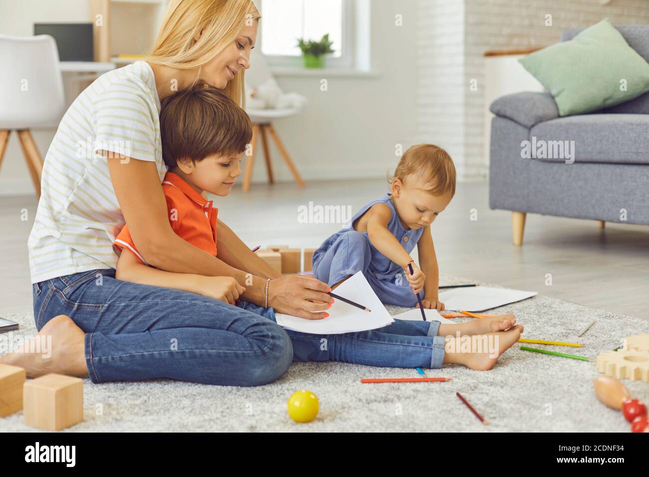 Die junge Mutter lehrt ihre beiden kleinen Kinder, mit Buntstiften zu zeichnen, während sie im Zimmer sitzt. Stockfoto