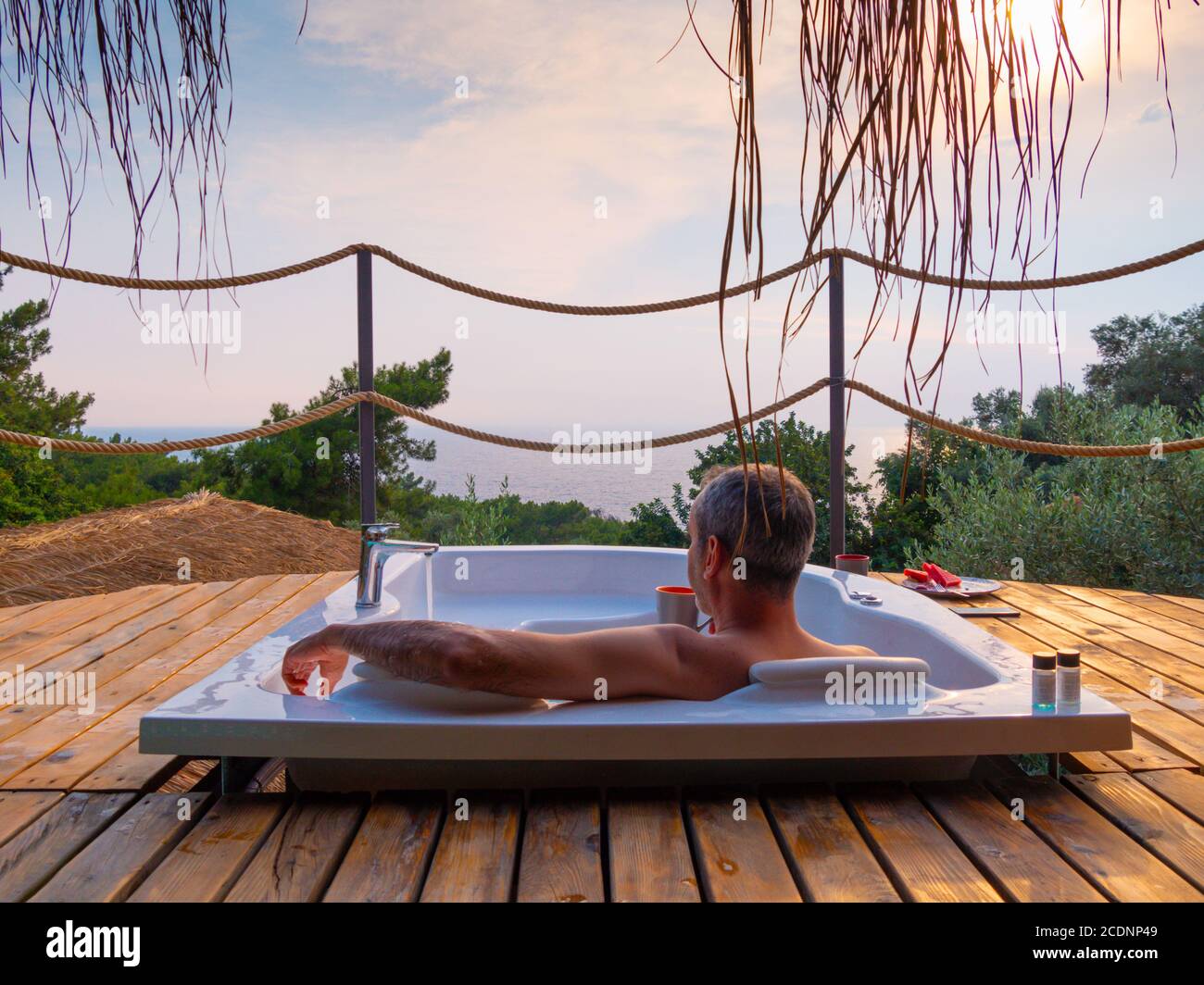 Ein hübscher erwachsener Mann liegt gekühlt in einem Whirlpool auf einem luxuriösen exotischen Spa Resort Hotelbalkon mit Meer und Sonnenuntergang. Stockfoto