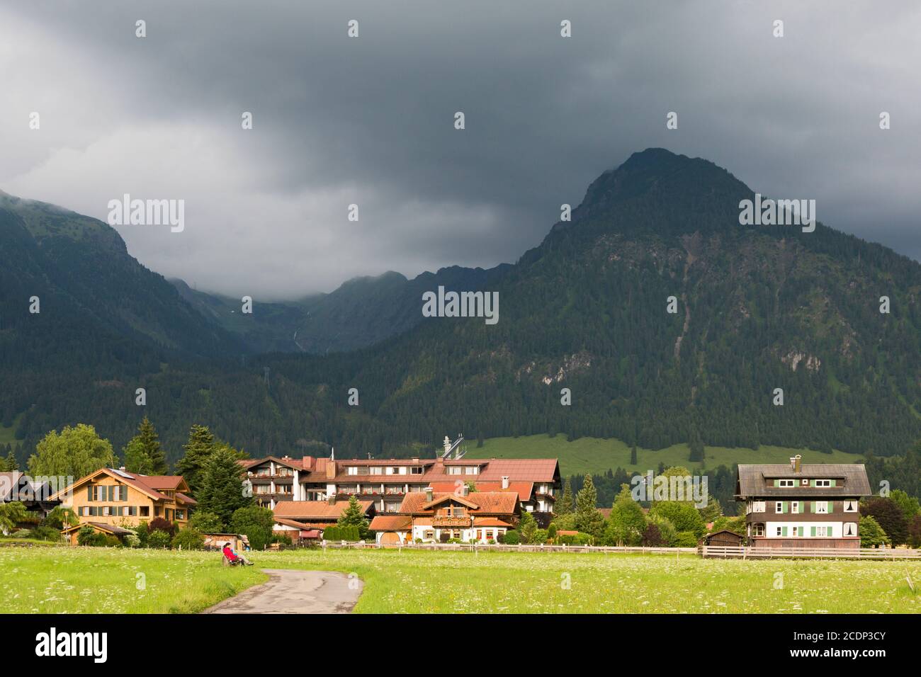 Paar auf einer Bank, Wiesenweg, Sonne, Häuser vor bewaldeten Bergen, tiefliegende Wolken Stockfoto