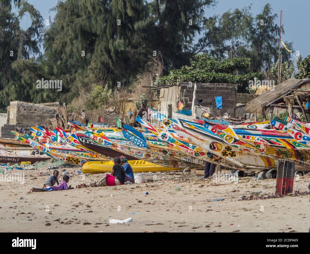 Nianing, Senegal - 24. Januar 2019: Afrikanische Familie sitzt in der Nähe der bunten hölzernen Fischerboote stehen am Sandstrand in Senegal. Afrika Stockfoto