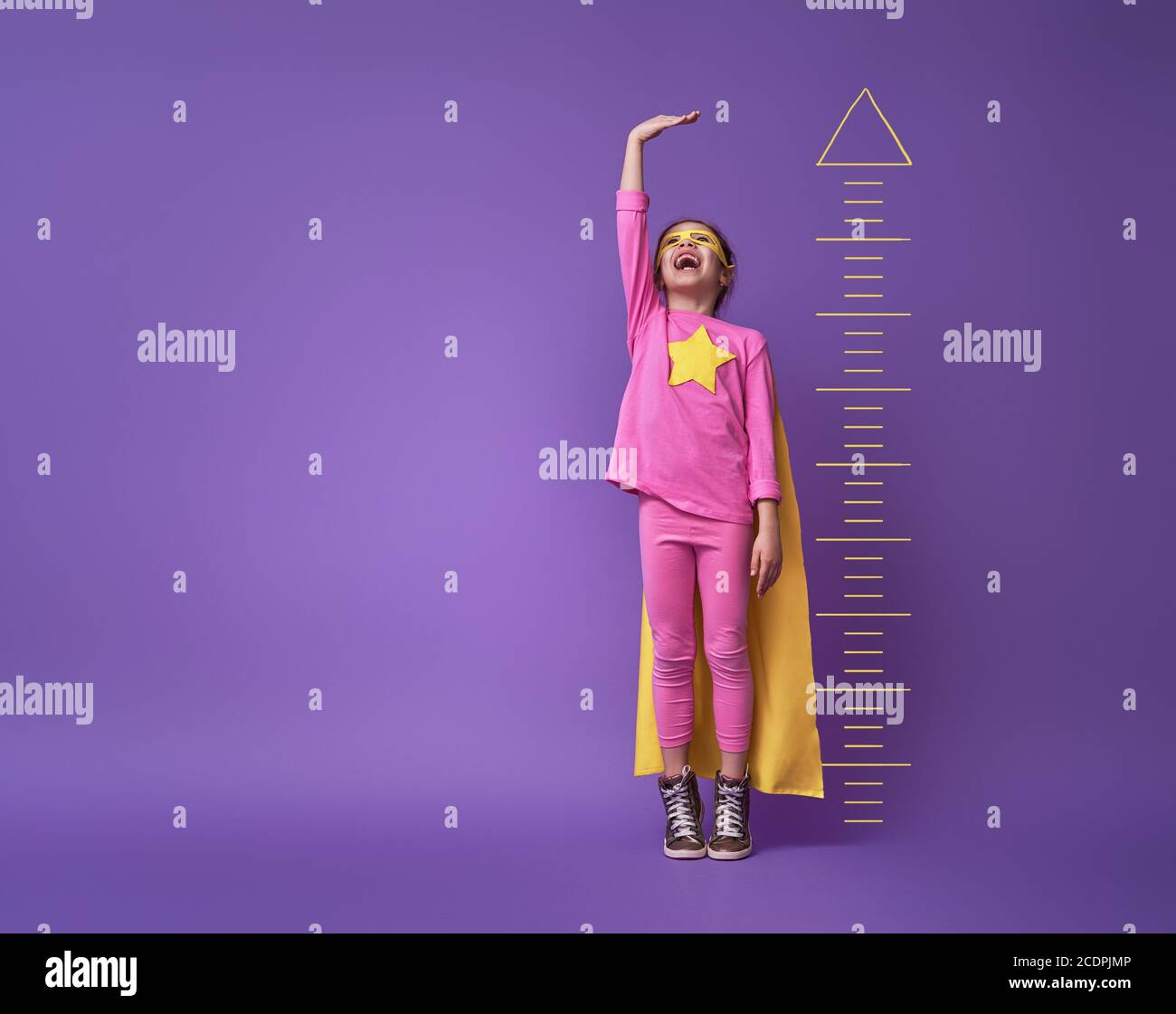 Kleines Kind spielt Superheld. Kid misst das Wachstum auf dem Hintergrund der hellen ultravioletten Wand. Girl Power-Konzept. Gelb, Rosa und pur Stockfoto