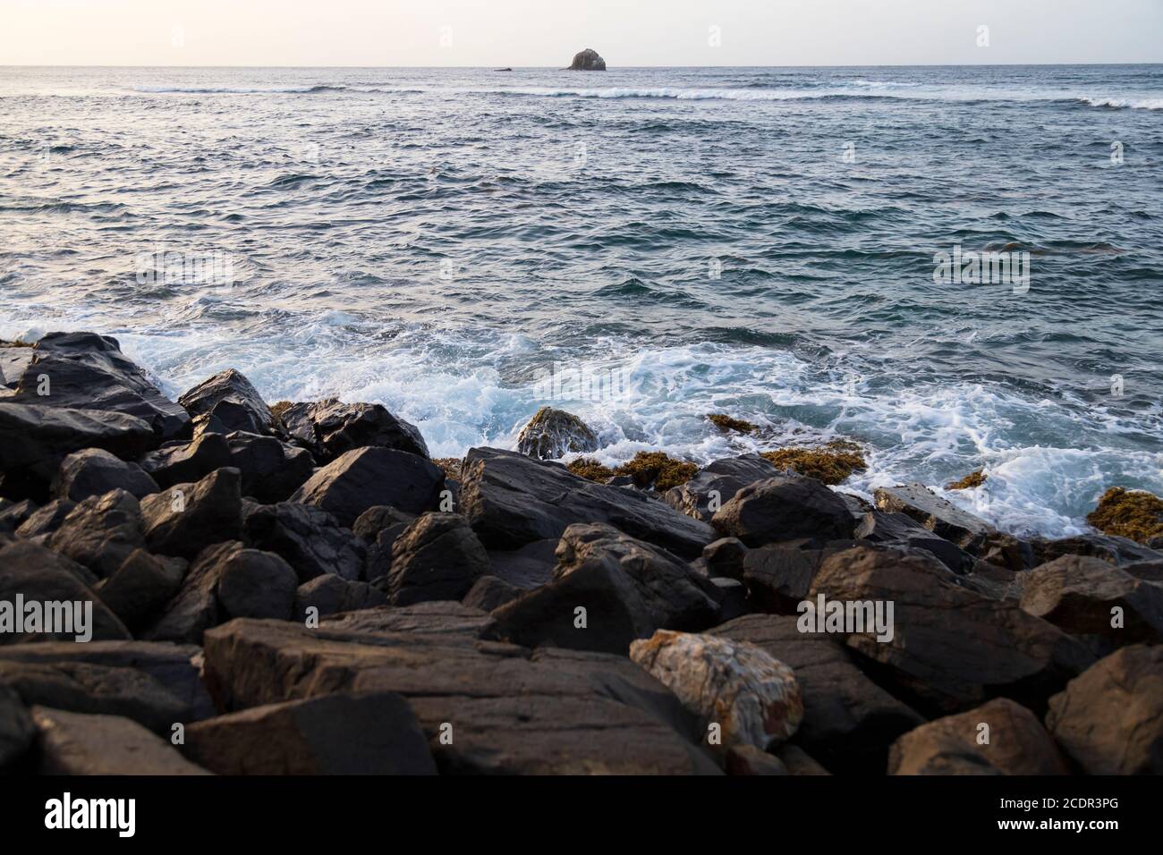 Eine Felsbarriere verwittert durch unerbittliche Wellenbewegung und ein Riesiger Felsblock auf dem Meer im fernen Hintergrund Stockfoto
