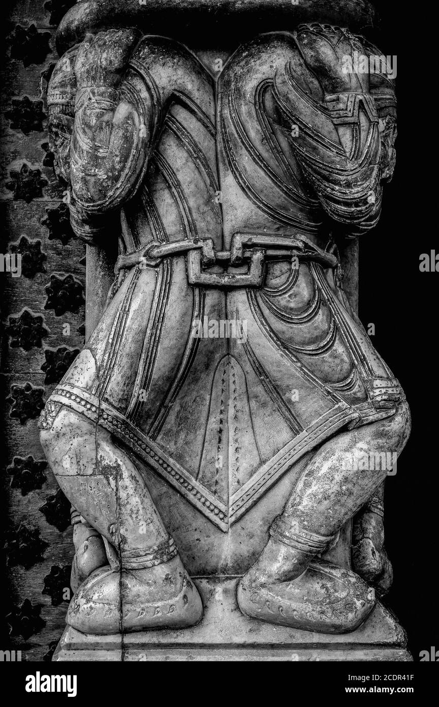 Gefesselter Gefangener trägt eine schwere Last. Mittelalterliche Steinkaryatid, die dazu beiträgt, ein gewichtiges romanisches Tympanum über dem westlichen Portal einer ehemaligen Kathedrale, heute die Elglise Sainte-Marie, in Oloron-Sainte-Marie, Nouvelle-Aquitaine, Frankreich, zu halten. Diese Karibik ist einer von zwei gefesselten Gefangenen, die an der Taille aneinander gekettet sind. Stockfoto