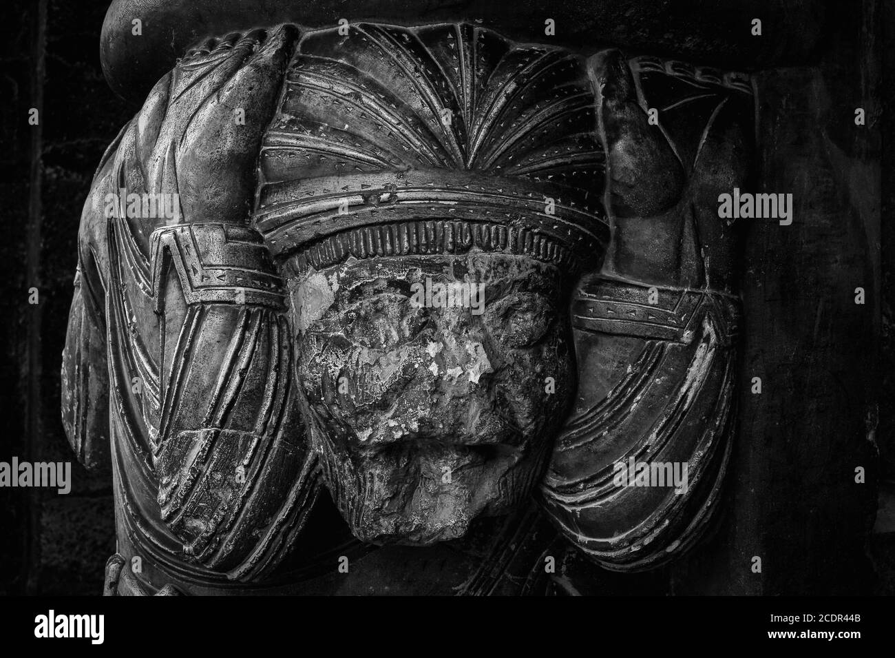 Das Gesicht eines erschöpften Mannes, der eine schwere Last trägt. Mittelalterliche Steinkaryatid, die hilft, ein schweres romanisches Tympanum in einer französischen Kirche zu halten. Über dem Westportal der Ellen Sainte-Marie in Oloron-Sainte-Marie, Nouvelle-Aquitaine, Frankreich. Diese Karikatid ist eine von zwei - gefesselte Gefangene, an der Taille angekettet. Stockfoto
