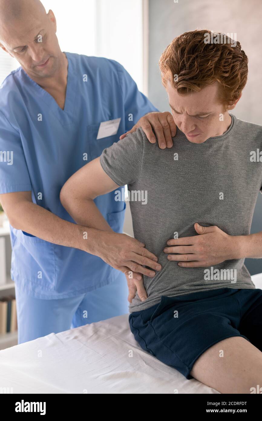 Junger Mann, der dem Arzt zeigt, wo es weh tut, und ihn berührt Zurück in Kliniken Stockfoto
