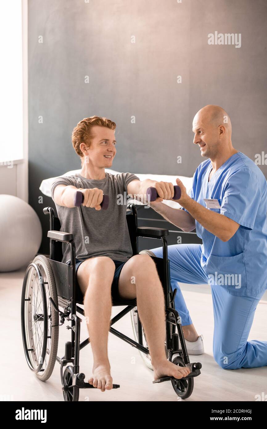 Reifer Arzt in Uniform hilft jungen Patienten mit Kurzhanteln in Rollstuhl Stockfoto