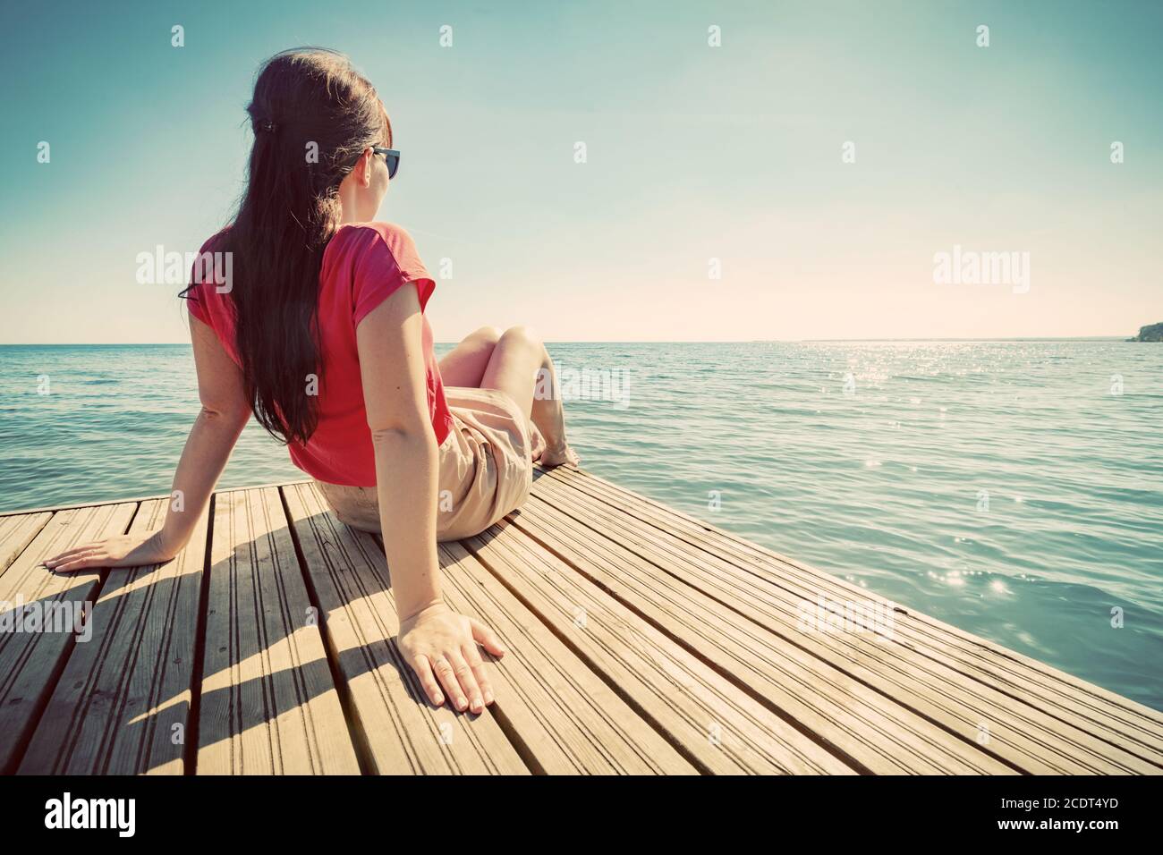 Junge Frau, die sich am Steg ausruhte und am sonnigen Sommertag das ruhige Meer betrat. Stockfoto