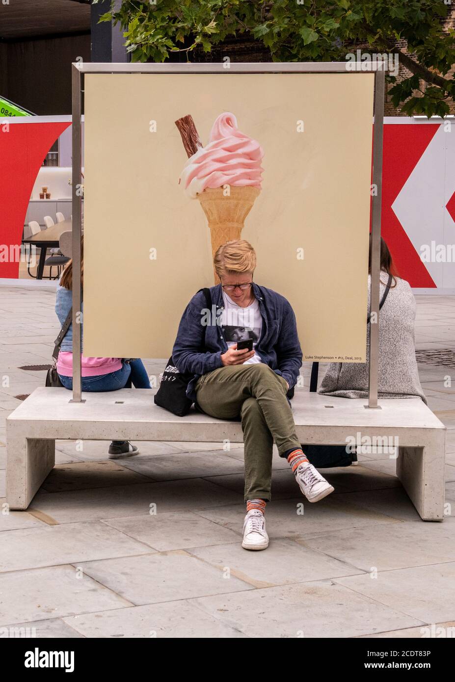 Junger Erwachsener sitzt auf der Bank mit Bild von Eis Kegel, Kings Cross, London, England Stockfoto