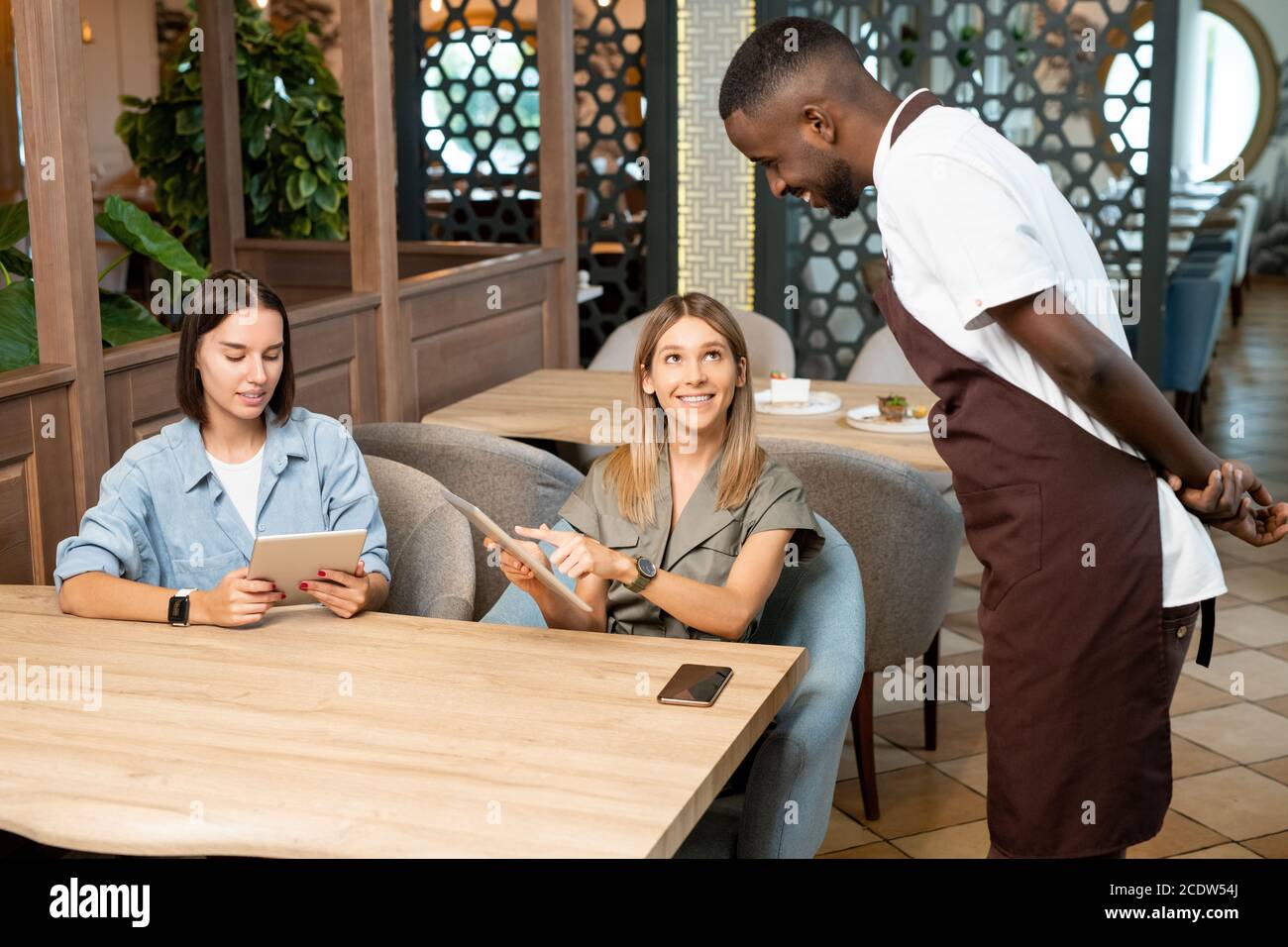 Junger lächelnder Kellner in Uniform, der sich über den Tisch beugte und schaute Online-Menü Stockfoto