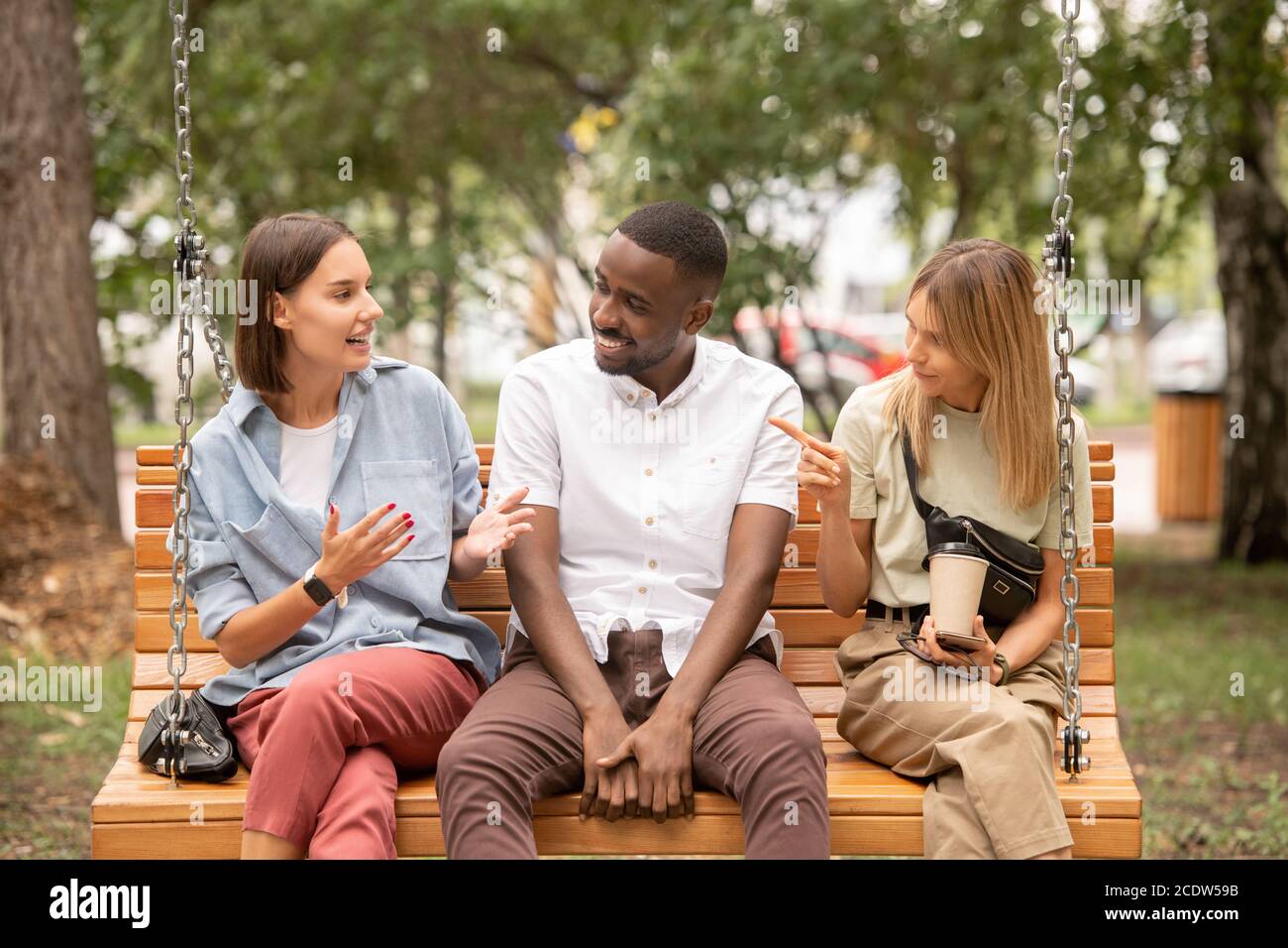 Glücklicher junger afrikanischer Mann, der auf hölzerner Schaukel zwischen zwei sitzt Streitenden Mädchen Stockfoto