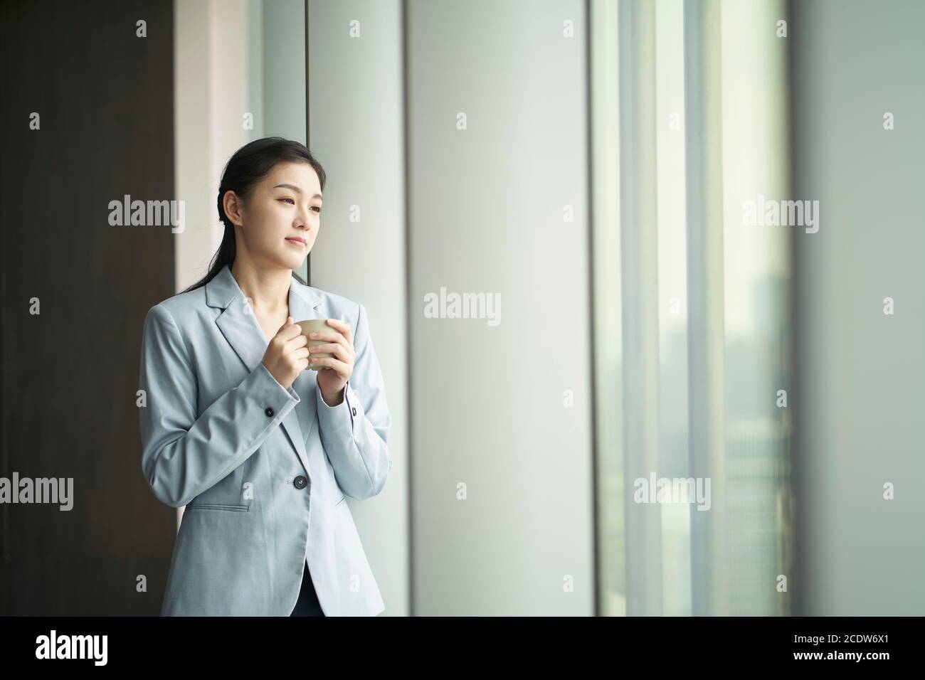 Junge asiatische Geschäftsfrau steht am Fenster mit einer Tasse Von Kaffee, der aussicht Stockfoto