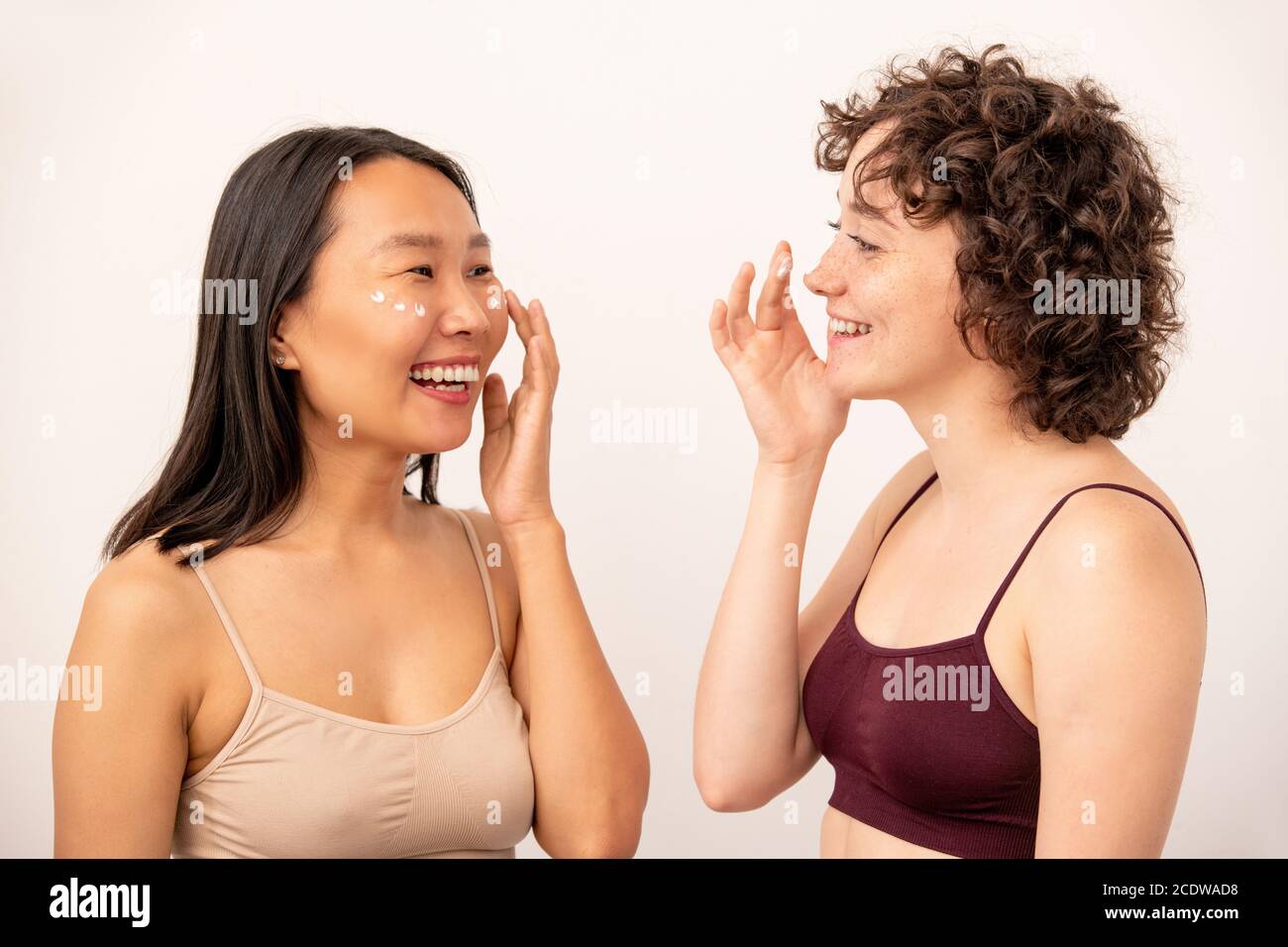 Zwei junge lachende interkulturelle Frauen in Tanktops Anwendung verjüngende Creme Stockfoto