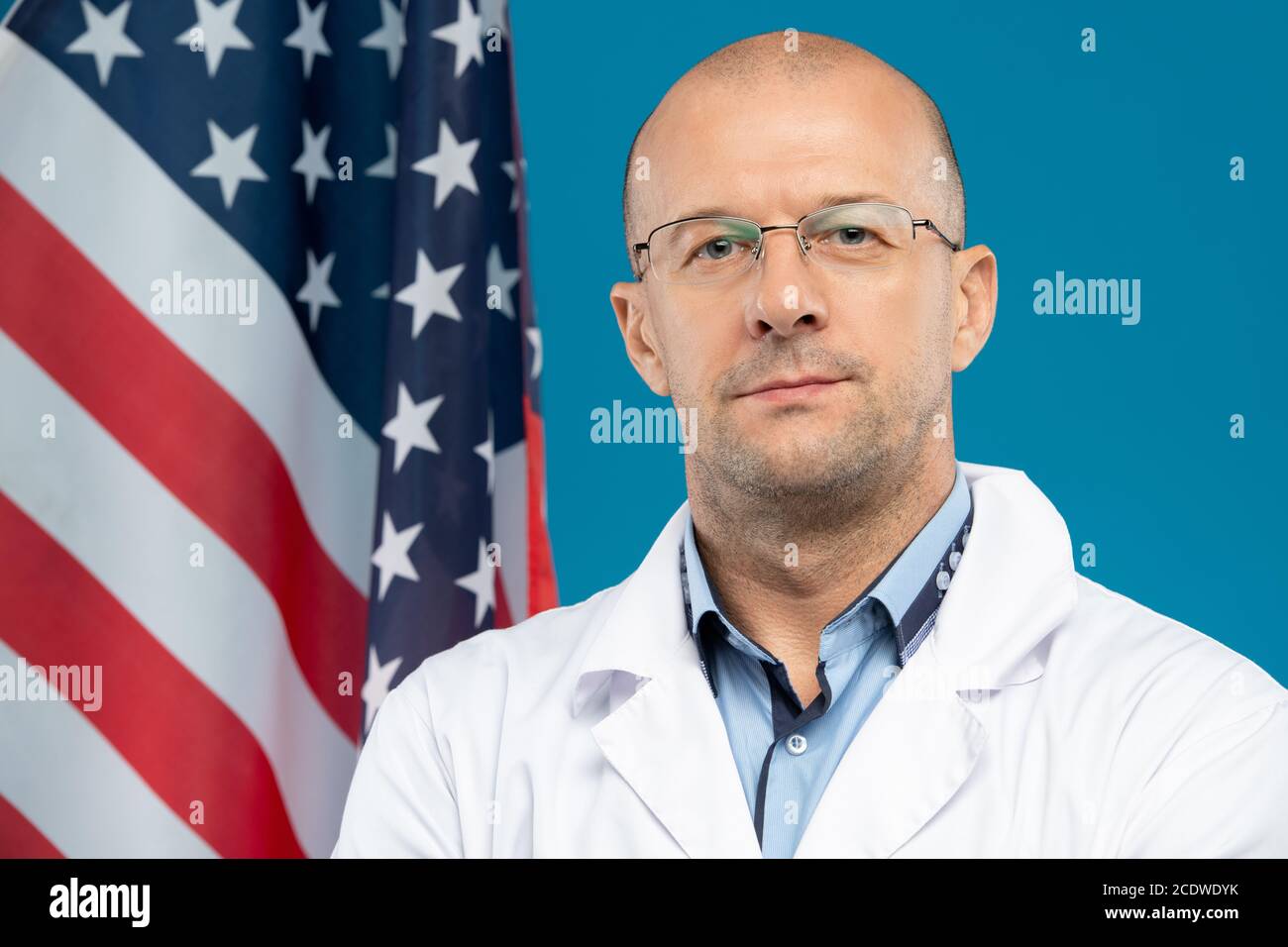 Glatzköpfiger Arzt mittleren Alters in Brillen und Weißkopfflächen, die dagegen stehen US-Flagge Stockfoto