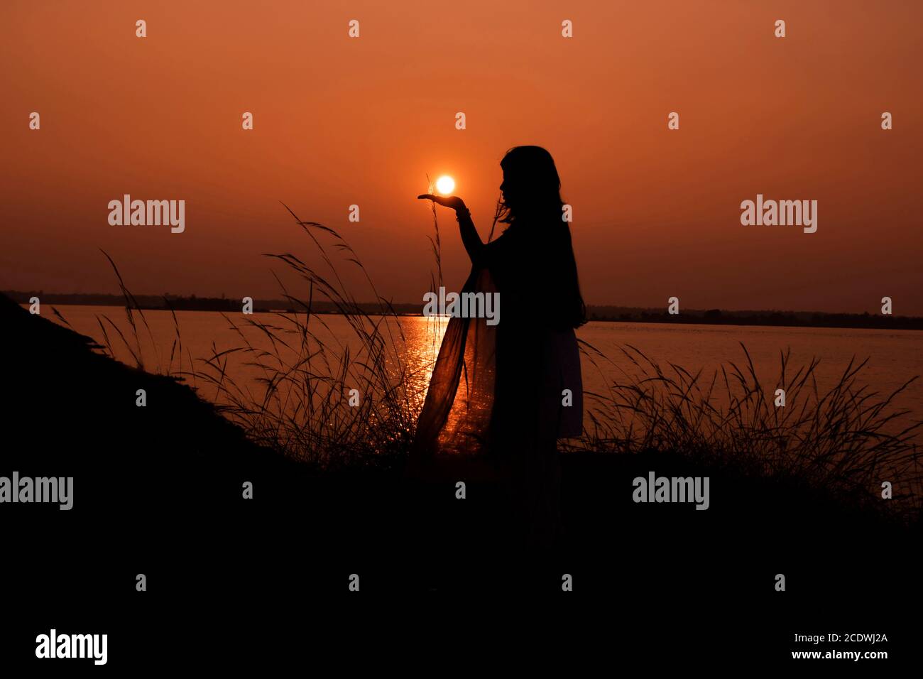 Am 5. Januar 2020 orange Sonnenuntergang hinter Gras Silhouette Mädchen Frau hält Sonne auf der Hand in Bakerswar Damm, birbhum, West Bengal, Indien Stockfoto