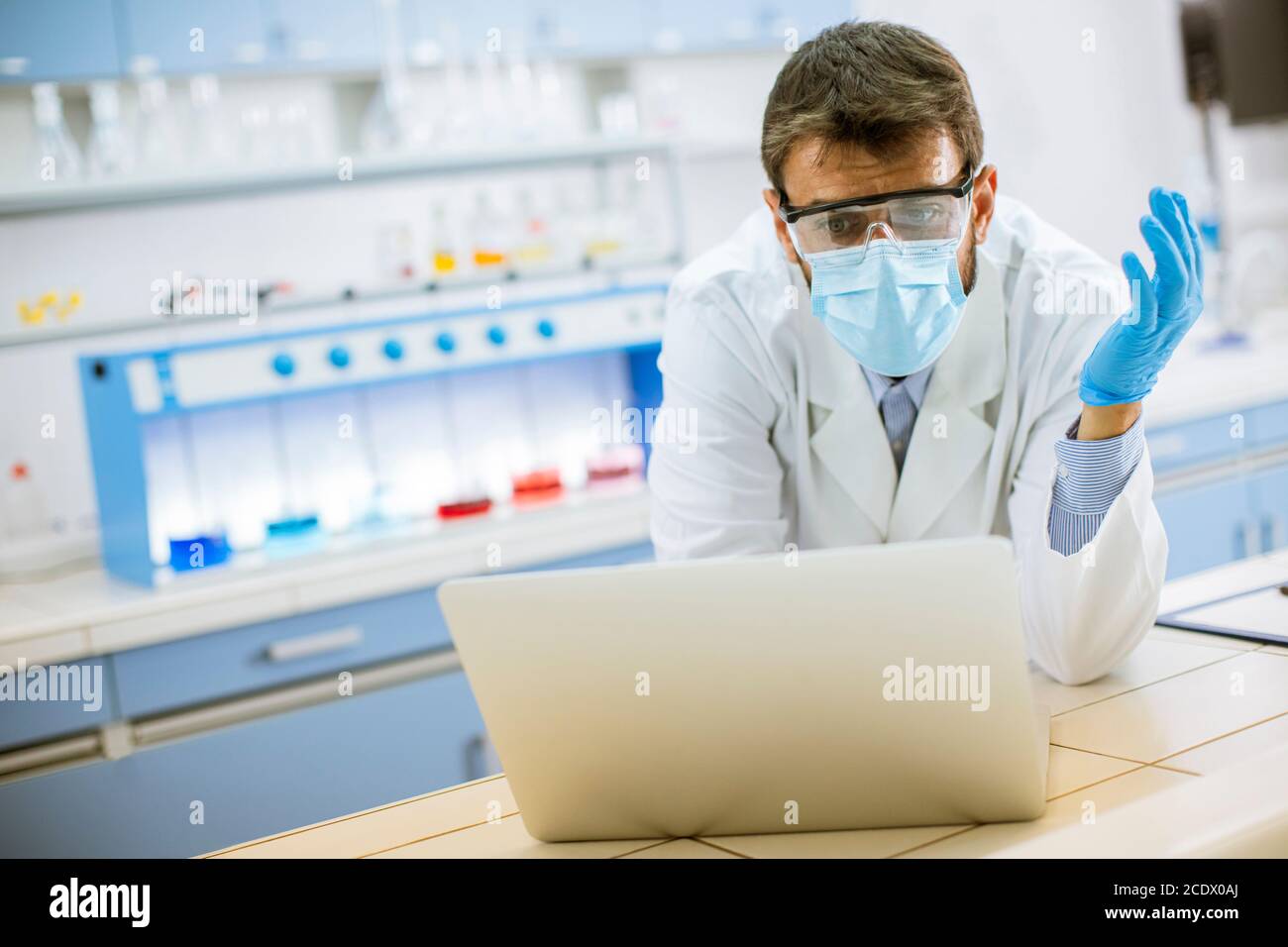 Hübscher junger Forscher in weißem Labormantel und Schutzmaske Arbeiten mit einem Laptop im Labor Stockfoto