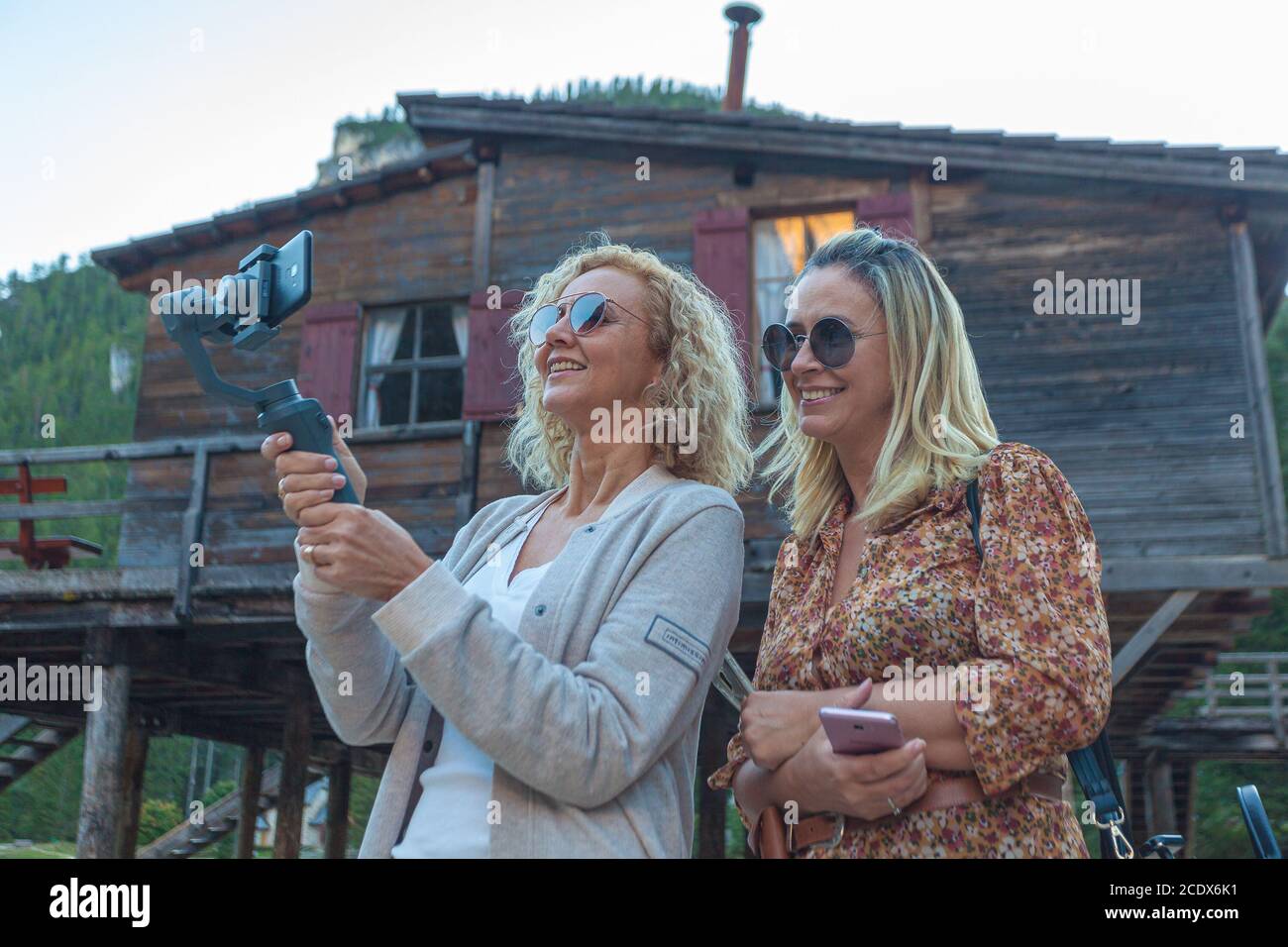 Paar brasilianische blonde Frauen beobachten Telefon mit Haus des Sees Prags Hintergrund, Südtirol, Italien Stockfoto