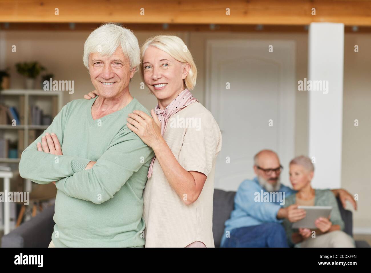 Porträt eines glücklichen älteren Paares, das im Stehen die Kamera anlächelt Im Raum mit einem anderen Paar im Hintergrund Stockfoto
