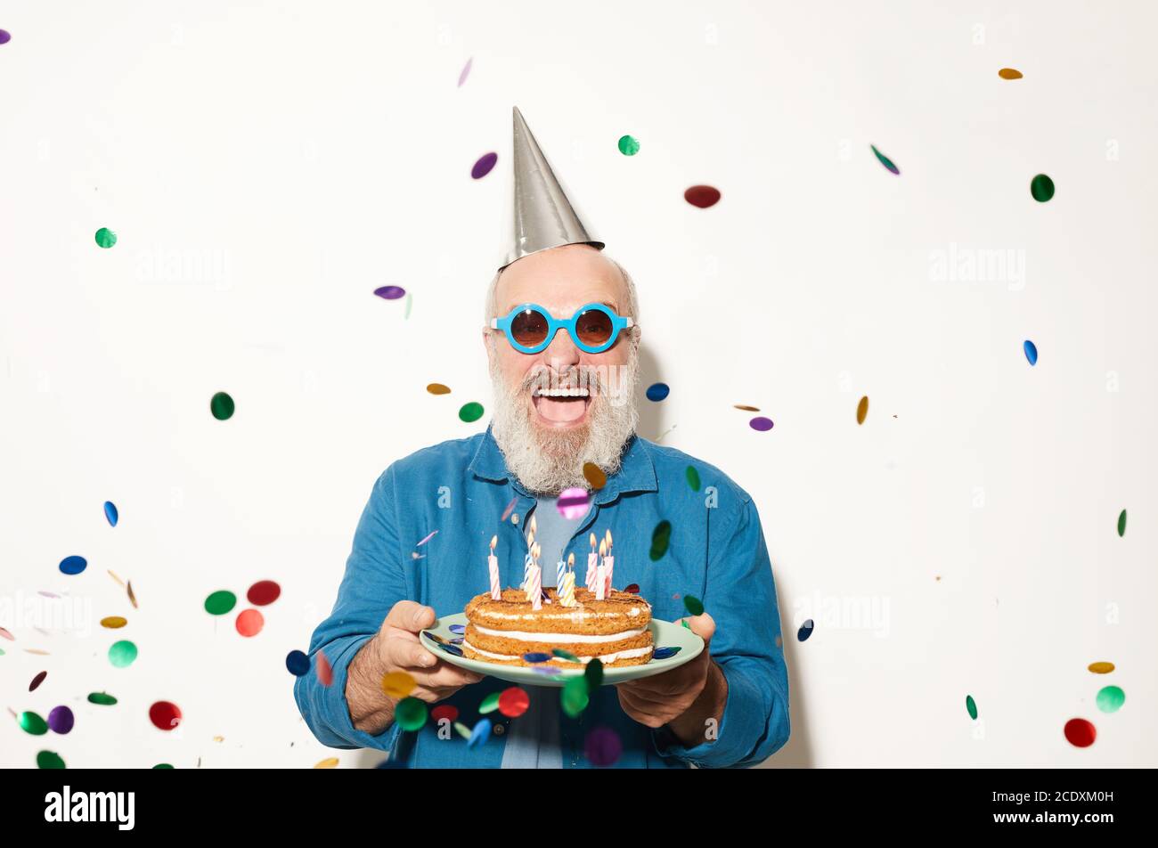 Porträt eines aufgeregten älteren Mannes in Sonnenbrillen, der die Kamera anlächelt Unter dem Konfetti stehen und Kuchen isoliert halten Weißer Hintergrund Stockfoto