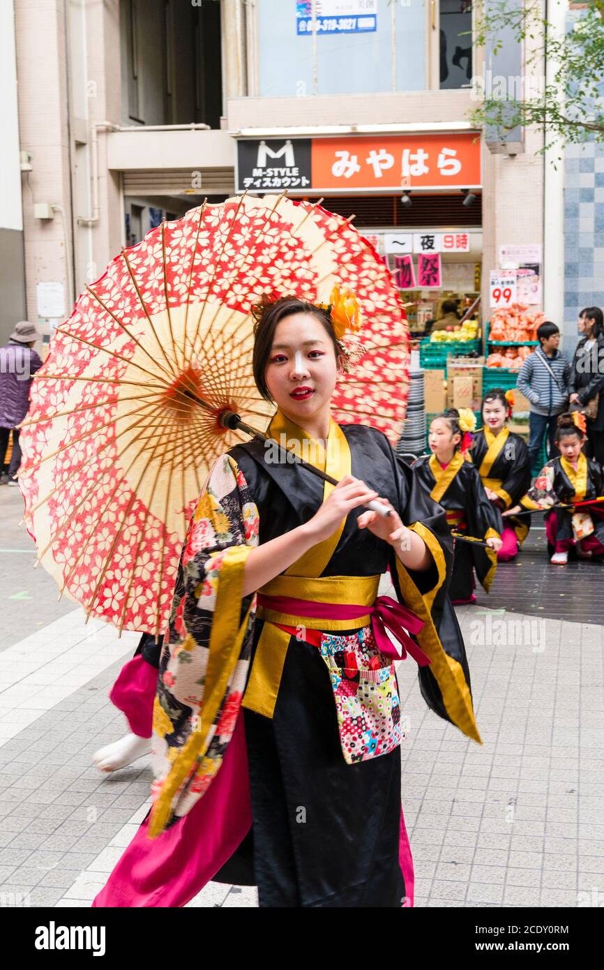 Nahaufnahme einer japanischen Frau, die Teil eines yosakoi-Tanzteams ist, die tanzt, während sie einen Sonnenschirm hält und direkt auf den Zuschauer schaut. Kyusyu Gassai Festival. Stockfoto